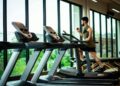 Young man workouts on treadmill in modern gym with large windows and natural light.