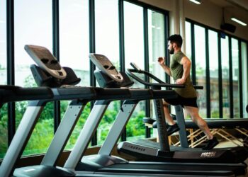 Young man workouts on treadmill in modern gym with large windows and natural light.
