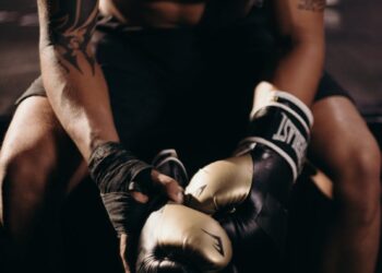 A muscular boxer with tattoos sitting shirtless in a gym holding boxing gloves.