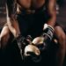 A muscular boxer with tattoos sitting shirtless in a gym holding boxing gloves.