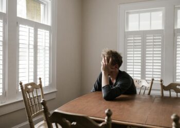 A man sits alone at a table in a bright room, displaying deep contemplation.